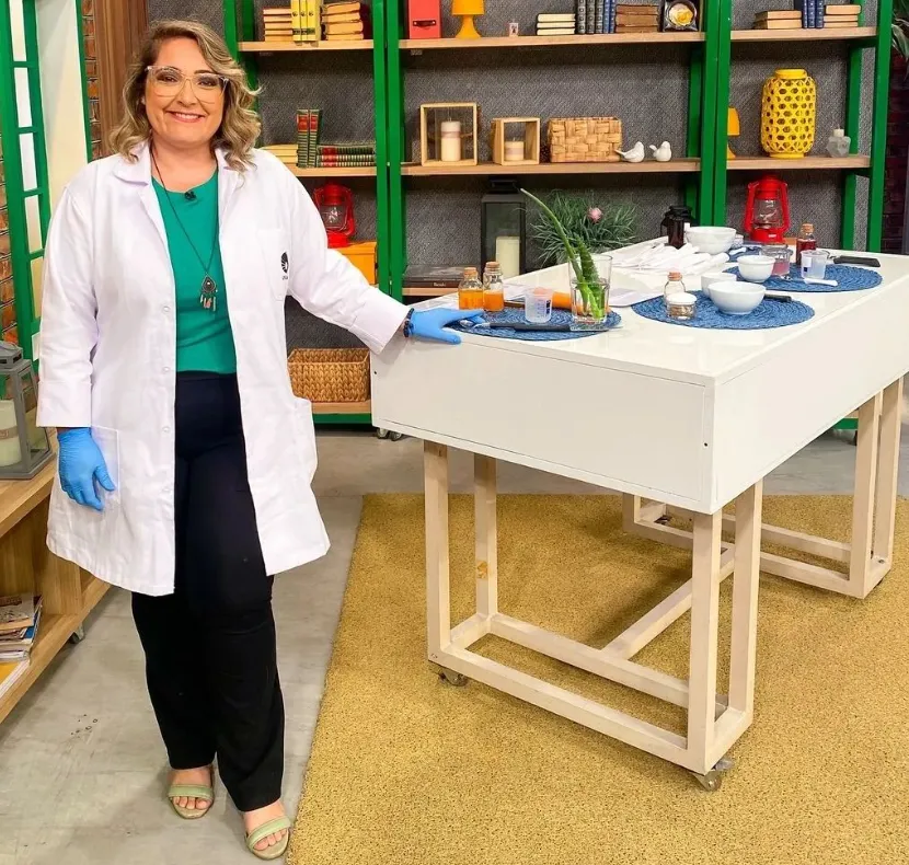 Imagem mostra a quimica Fernanda Honorato ao lado de uma mesa com óleos essenciais, plantas e flores aromáticos utilizados para fazer perfumes naturais